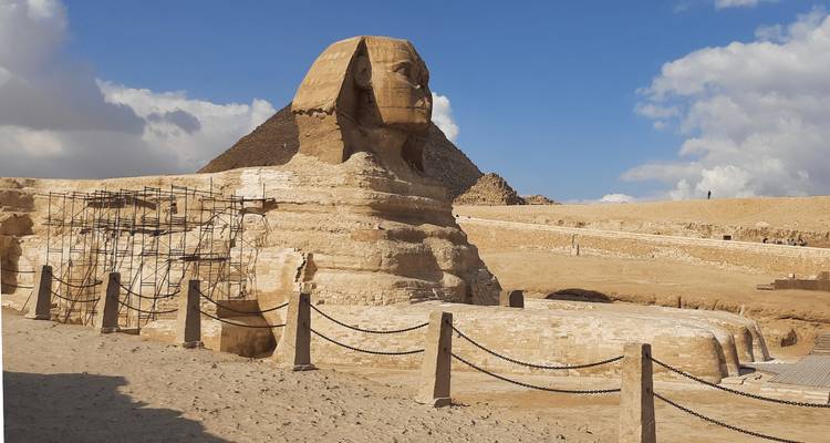 La Gran Esfinge se alza ante un cielo parcialmente nublado, su cuerpo de piedra caliza erosionado pero imponente junto a las pirámides.