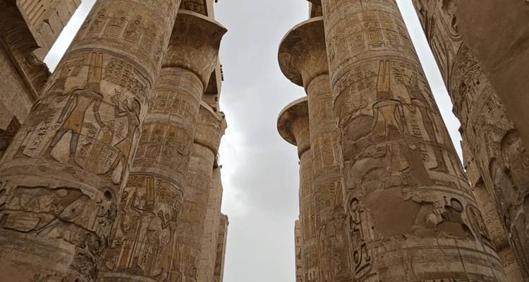 Vista hacia arriba de colosales columnas de piedra talladas con jeroglíficos en Karnak se alzan contra un cielo gris.