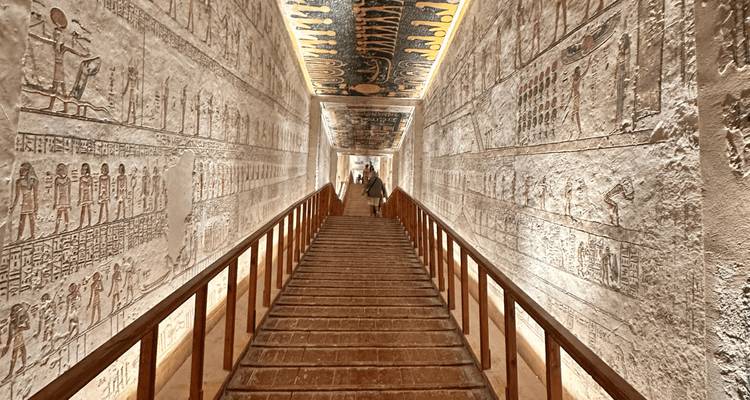 Paredes con jeroglíficos iluminados flanquean una rampa de madera que conduce profundamente hacia un corredor de tumba egipcia ornamentalmente decorado.