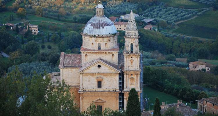 Église en pierre de style Renaissance avec un grand dôme et un clocher attenant située parmi les cyprès, les oliviers et les collines toscanes en patchwork.