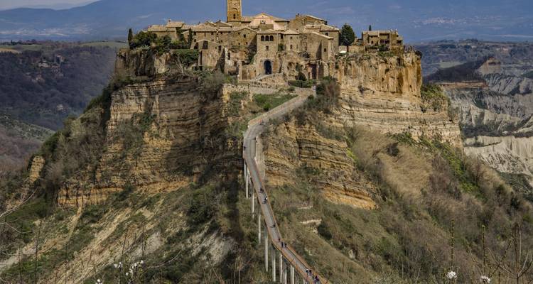 Village spectaculaire au sommet d'une falaise accessible par un long pont piétonnier, entouré de ravins profonds et de montagnes lointaines.