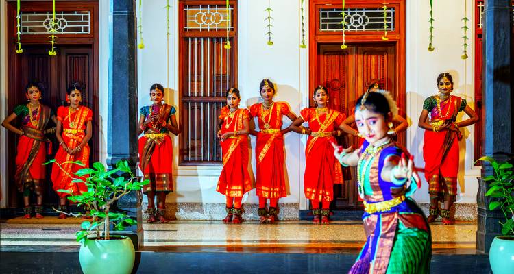 Traditional Bharatanatyam dancers in vibrant red costumes perform inside a heritage courtyard.