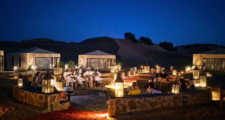 Evening scene of a luxury desert camp with guests seated around a fire pit and lanterns glowing against tall dunes.