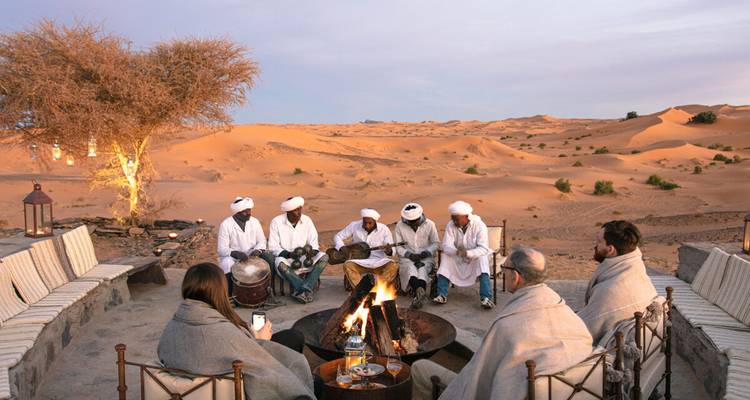 Local musicians perform around a campfire in the desert at dusk while guests sit wrapped in blankets.