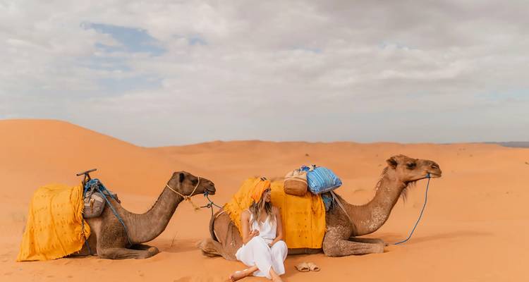 Woman sits beside two saddled camels resting on orange sand dunes under a partly cloudy sky.