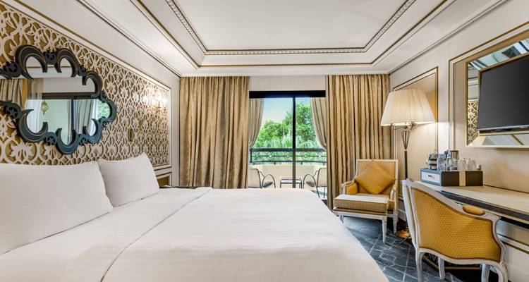 Classic hotel bedroom with patterned wallpaper, gold accents and a balcony view of greenery.