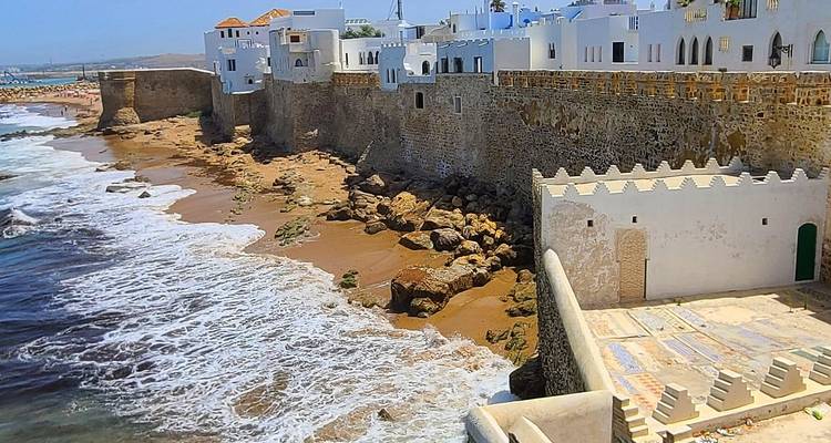 Whitewashed seafront ramparts of Asilah with waves crashing on the sandy shore below.