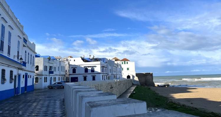 Row of blue-trimmed seaside houses in Asilah beside a breezy beach and stone walkway.