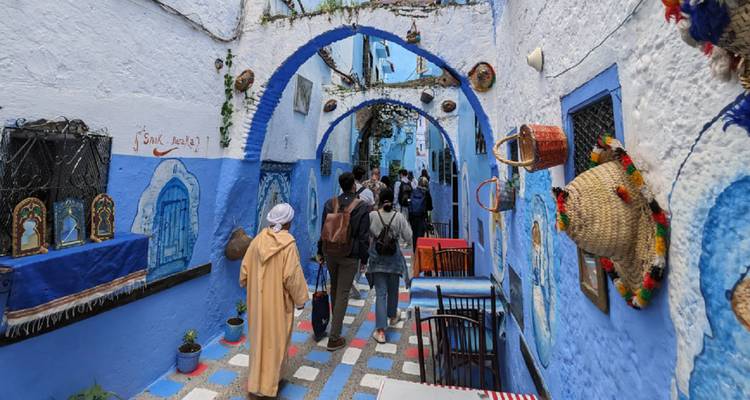 Tourists walk through a narrow blue-painted alley in Chefchaouen decorated with local crafts.