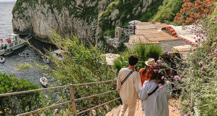 Turistas caminando por un sendero bordeado de flores hacia acantilados marinos escarpados y barcos atracados en Capri.