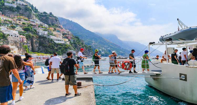 Los visitantes caminan por una pasarela para abordar un barco en un puerto soleado bajo casas empinadas y coloridas en el acantilado.