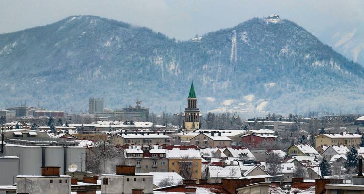 Snow-dusted cityscape with a green-topped church steeple set against forested hills blanketed in winter white.