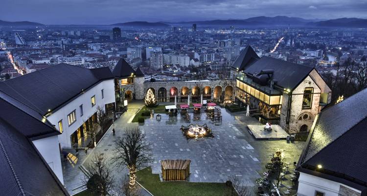 Evening lights illuminate Ljubljana Castle courtyard overlooking the twinkling city and distant mountains at dusk.