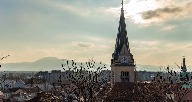 Tall clock-tower spire rises above rooftops against a hazy sunset sky and distant mountains.
