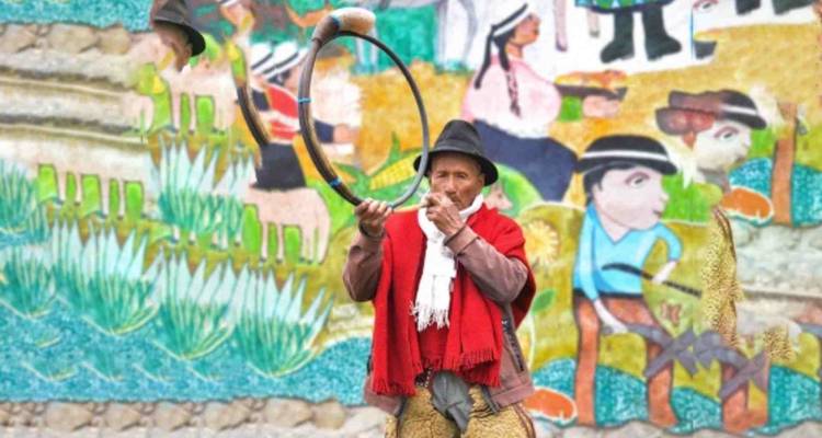 Intérprete indígena con vestimenta tradicional tocando un instrumento circular con un fondo de mural colorido.
