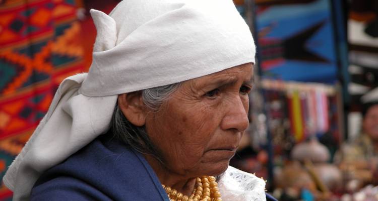 Retrato de una mujer indígena mayor usando un pañuelo blanco en la cabeza y collares de cuentas doradas en capas contra un fondo de mercado.