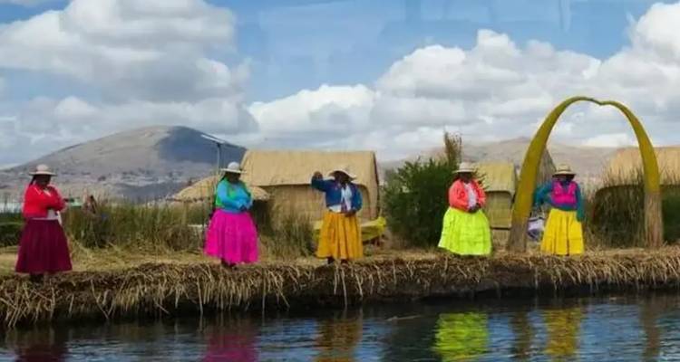 Frauen in bunten traditionellen Röcken stehen auf einer Schilfsinsel neben ruhigem Wasser und Bergen.