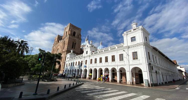 Escena callejera de la época colonial en Cuenca con edificio blanco con arcadas, torre de piedra y cúpulas de catedral bajo un cielo azul con nubes dispersas