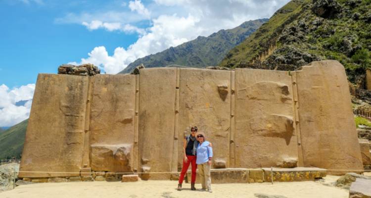 Dos turistas posando frente a un enorme muro de piedra tallada en un sitio arqueológico inca bajo picos montañosos