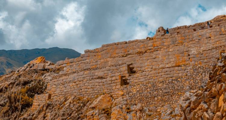 Largo muro de piedra inca aterrazado que asciende por una ladera bajo nubes dramáticas y cielo azul