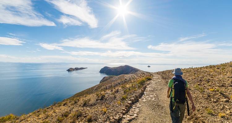 A hiker walks a cliff-top trail overlooking the deep blue waters of Lake Titicaca under bright sun.