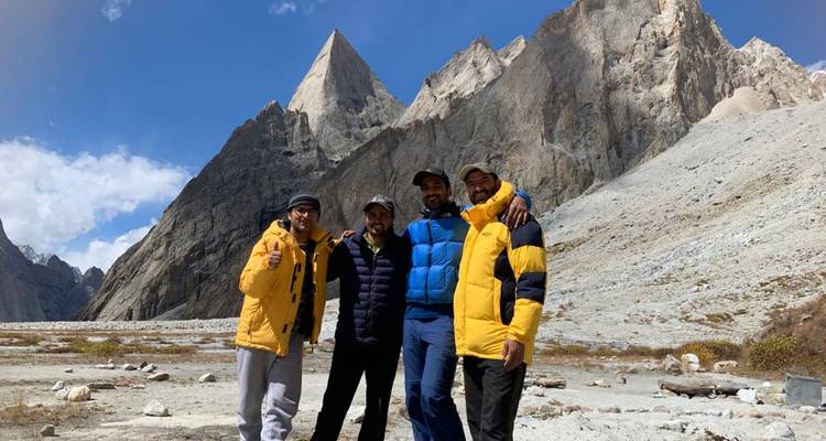 Vier trekkers in geïsoleerde jassen poseren in een kale alpine vallei met dramatische granieten spitsen op de achtergrond onder een blauwe hemel.