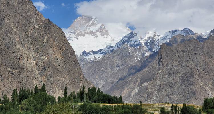 Torenhoge ruige bergen met besneeuwde toppen rijzen op boven groene populierenbossen onder een gedeeltelijk bewolkte hemel.