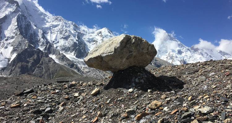 Grote gebalanceerde rotsblok op donker steenpuin met met sneeuw bedekte Karakoram-toppen en blauwe lucht.