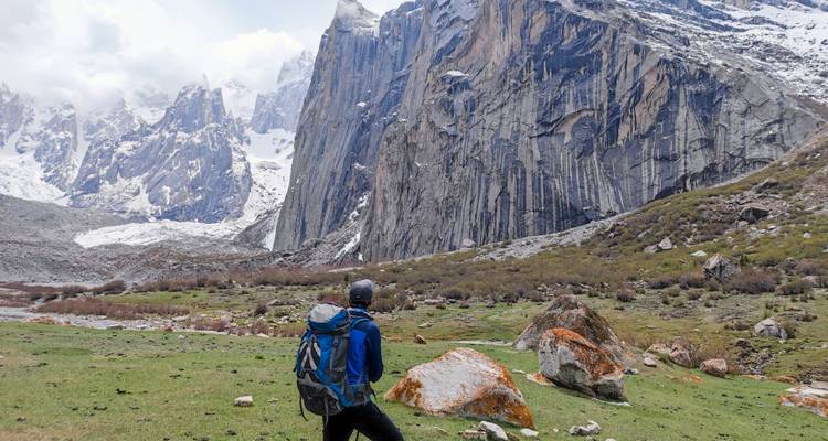 Eenzame rugzaktoerist kijkt naar torenhoge verticale granietrotsen en besneeuwde toppen over een groene alpenweidegrond.