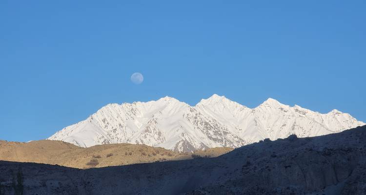 Besneeuwde bergkam onder een heldere blauwe lucht met een bleke volle maan die boven de horizon opkomt.