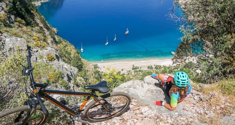 Cycliste se reposant au bord d'une falaise surplombant une baie turquoise et une plage de sable loin en contrebas.