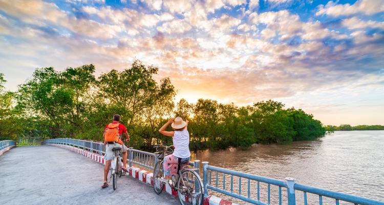 Twee fietsers pauzeren op een kleine brug over een serene rivier, bewonderend een kleurrijke zonsondergang en weelderige mangroves.