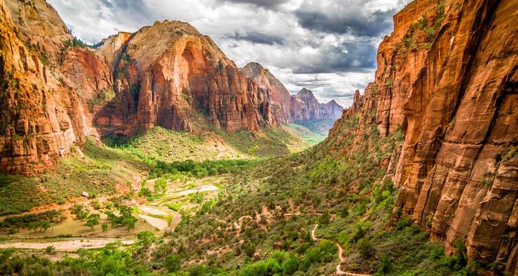 Dramatic view into Zion Canyon with winding trail and sunlit orange cliffs under moody clouds.