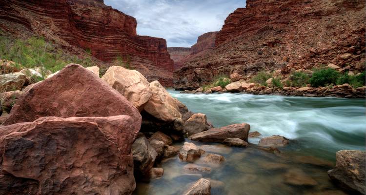 Reißender Fluss, der durch steile rote Canyonwände unter einem bewölkten Himmel fließt.