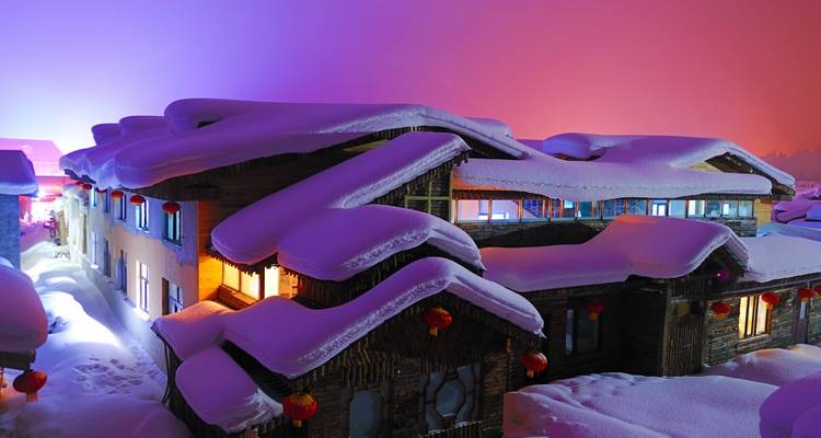 Snow-covered village houses glowing with warm lights beneath a colorful twilight sky.