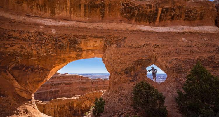 Un randonneur encadré dans une fenêtre de grès en forme de cœur contemple de vastes canyons de roche rouge.