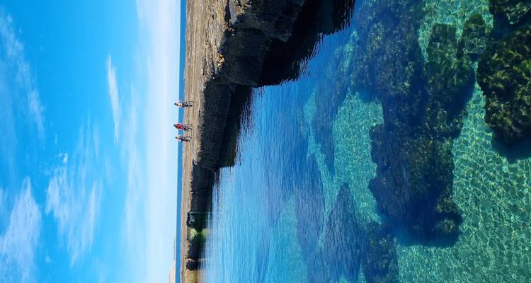 Piscine naturelle cristalline révélant des formations rocheuses tandis que des cyclistes roulent le long du rivage escarpé au-dessus.