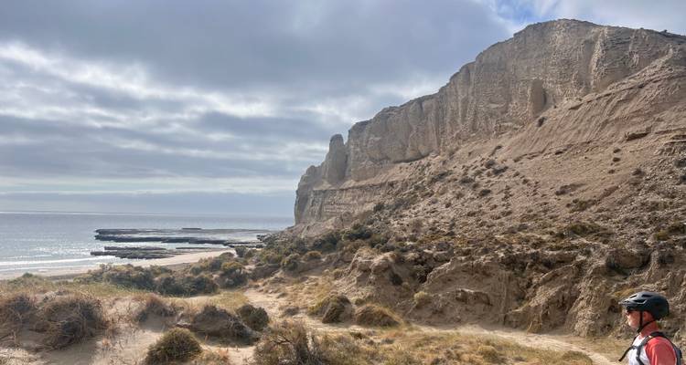 Cycliste de VTT faisant une pause à côté d'imposantes falaises marines beiges sous un ciel partiellement nuageux.