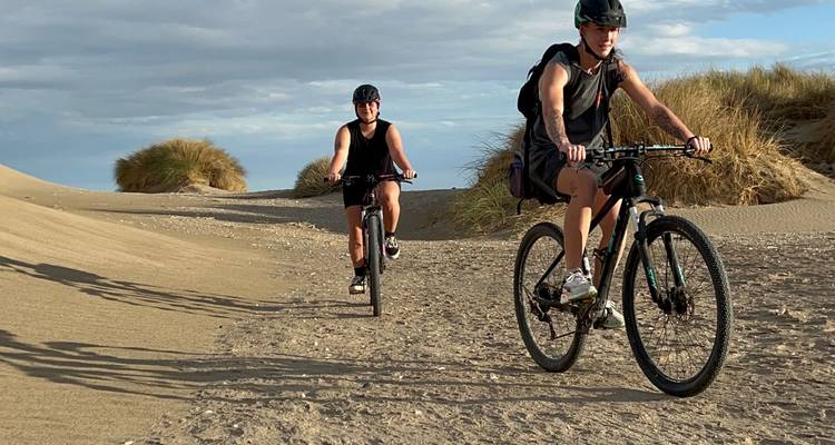 Deux cyclistes roulant à travers des dunes de sable avec des touffes d'herbe sous un ciel doux.