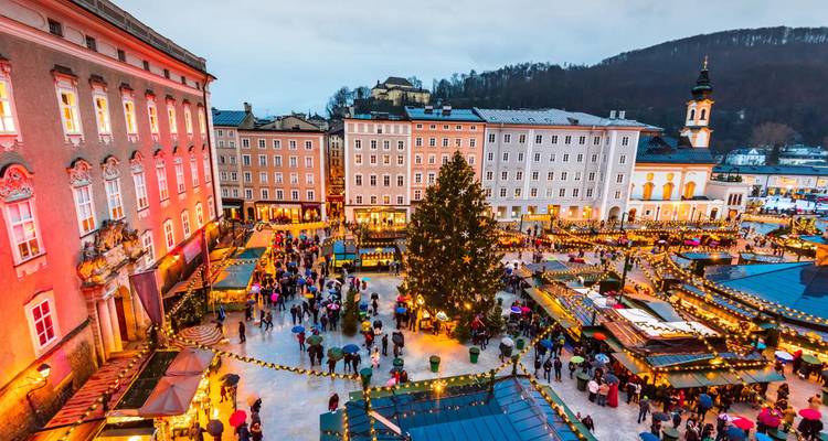 Ein lebendiger Weihnachtsmarkt erfüllt Salzburgs historischen Platz in der Dämmerung, glitzernde Lichter umgeben einen ragenden Baum.
