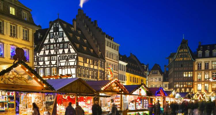 Fel verlichte houten vakwerkgebouwen en feestelijke kraampjes creëren een levendig kerstmarktscène op het blauwe uur in Straatsburg.