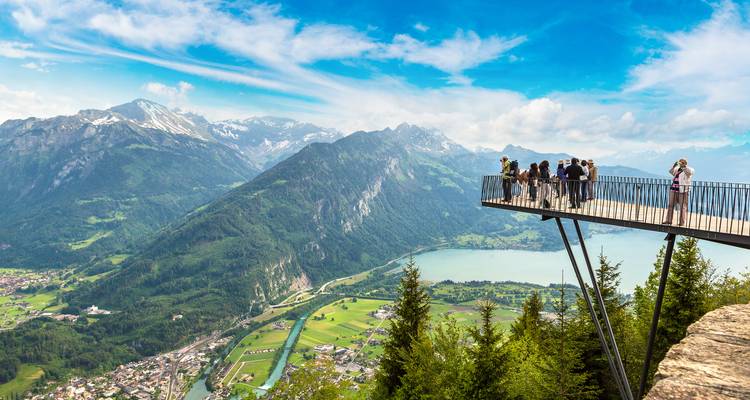 Visitors stand on a dramatic metal skywalk viewpoint above Interlaken with alpine lakes and peaks below.