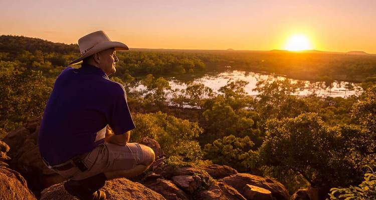 Un homme portant un chapeau de cow-boy est assis sur un point de vue rocheux, regardant un coucher de soleil orange éclatant au-dessus des plaines inondables et des zones boisées.