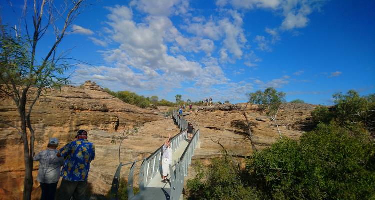 Les visiteurs traversent une passerelle d'acier enjambant des affleurements de grès rocheux sur fond de ciel bleu éclatant parsemé de nuages.