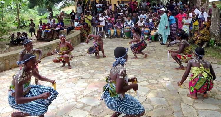 Danseurs traditionnels en tenues à motifs accroupis en formation devant un public rural attentif.