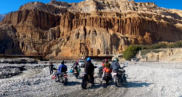 Des motocyclistes arrêtés sur un lit de rivière de gravier éclipsés par d'imposantes falaises de roche orange.