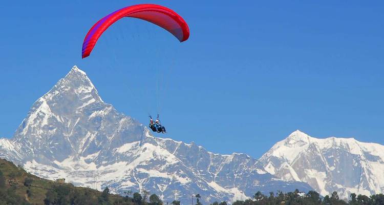 Un parapente biplace plane devant le sommet enneigé du Machhapuchhre sous un ciel bleu profond.
