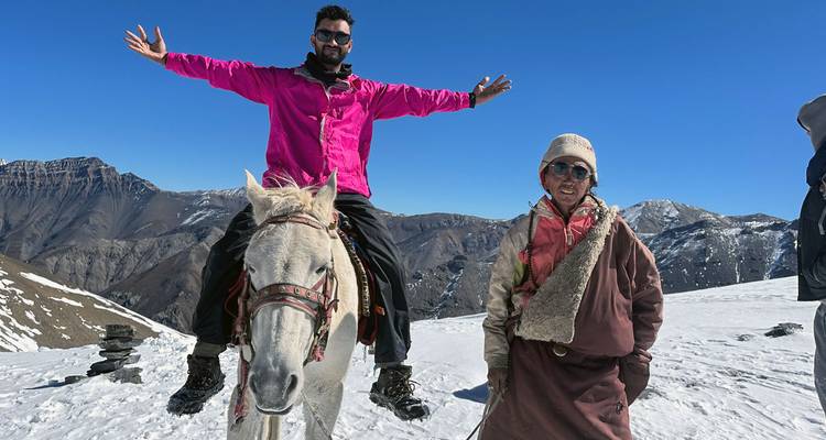 Reiziger steekt armen omhoog terwijl hij op een wit paard rijdt op een met sneeuw bedekte Himalaya-pas naast een plaatselijke gids.