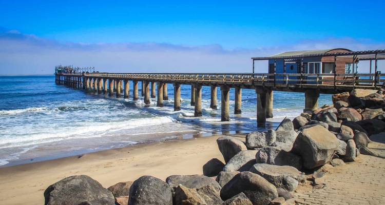 Long wooden jetty stretching over waves at Swakopmund beach under clear blue sky.
