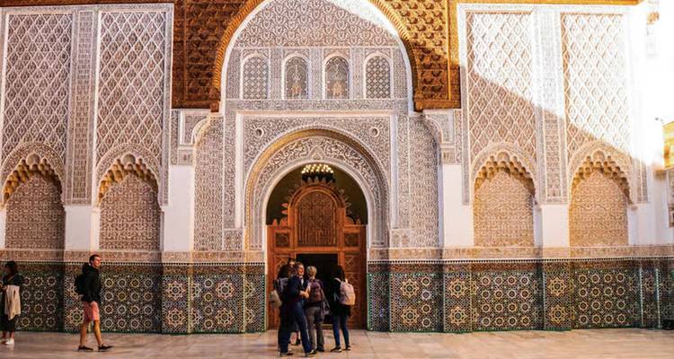 Fachada intrincadamente azulejada y entrada de cedro tallado de una madraza marroquí con visitantes admirando la artesanía